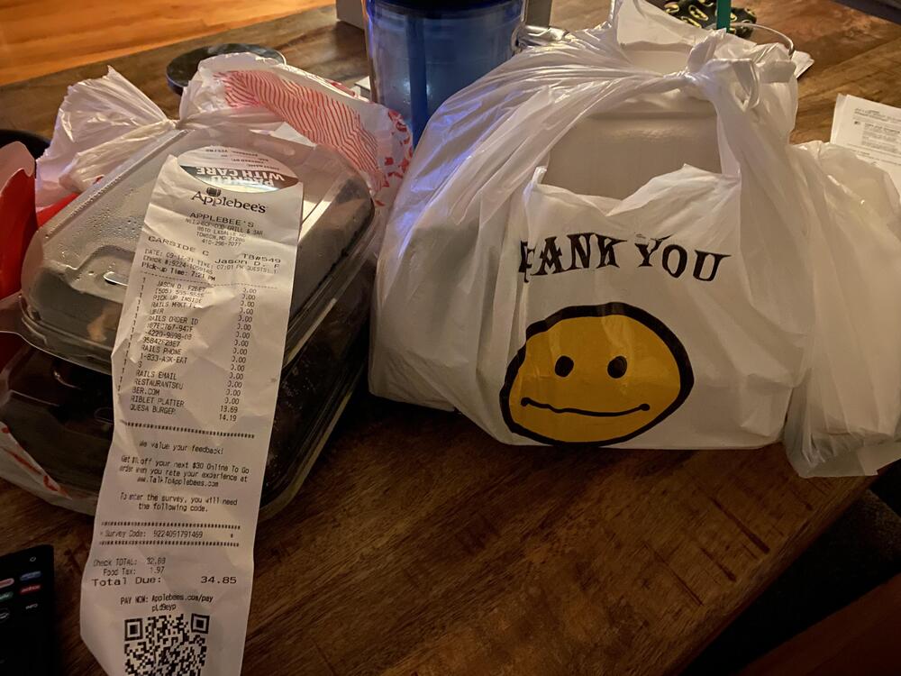 Takeout food in plastic bags sits on a table. One bag has a smiley face and “Thank You” printed on it. A long Applebee’s receipt is visible on top of the containers next to a blue drink cup.