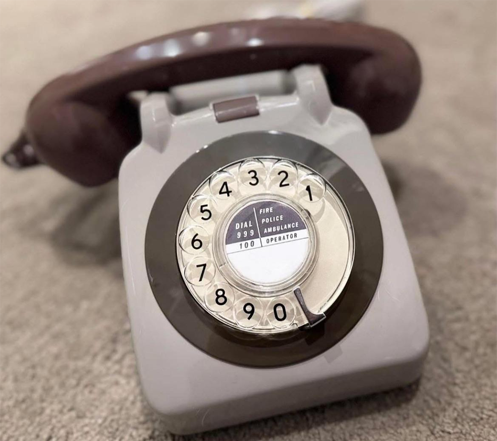 A vintage rotary dial telephone with a brown receiver and gray base, sitting on a carpeted surface. The dial lists emergency numbers for fire, police, ambulance, and operator.