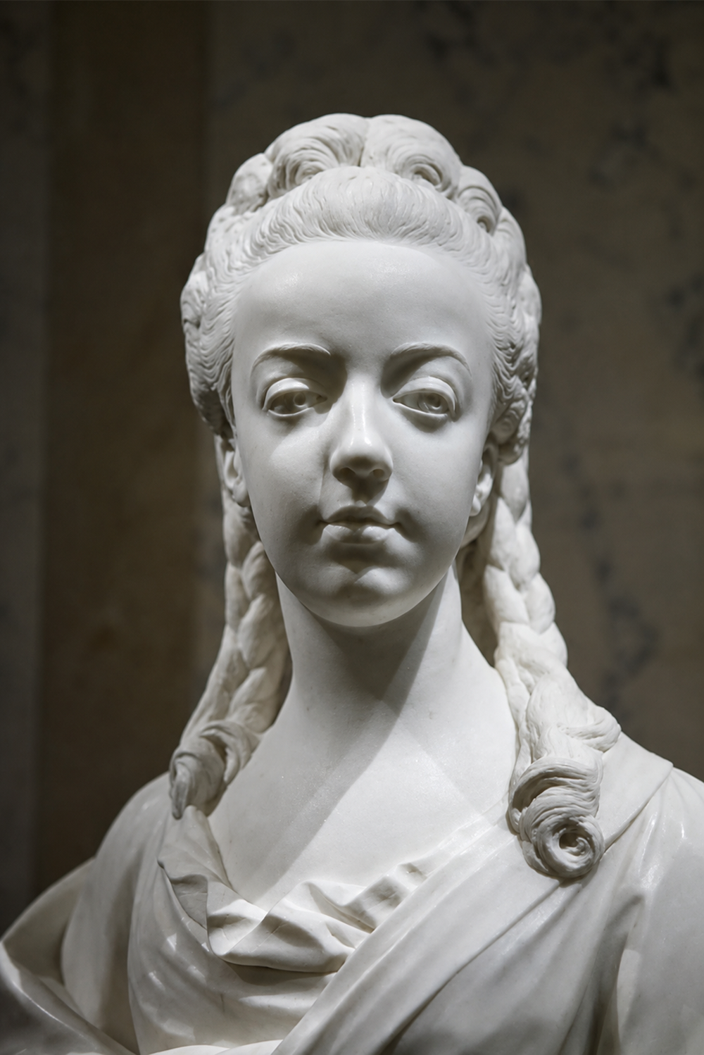 Marble bust of a young woman with braided hair and draped clothing, facing forward. The detailed sculpture highlights her delicate facial features and smooth texture.