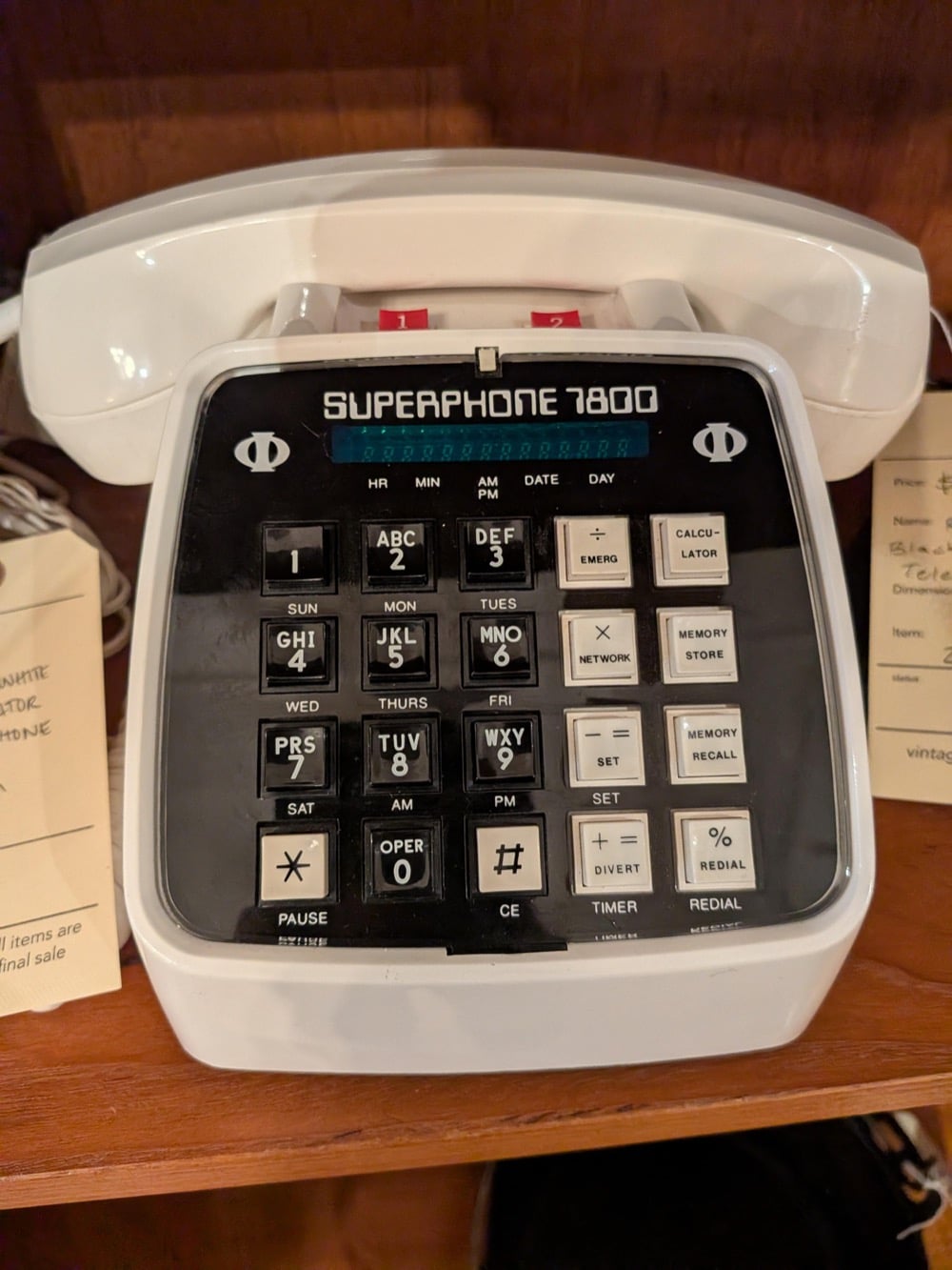 A white vintage Superphone 7800 sits on a wooden shelf, featuring a large keypad with numbers, symbols, and special function buttons. Two paper tags hang nearby, partially visible.