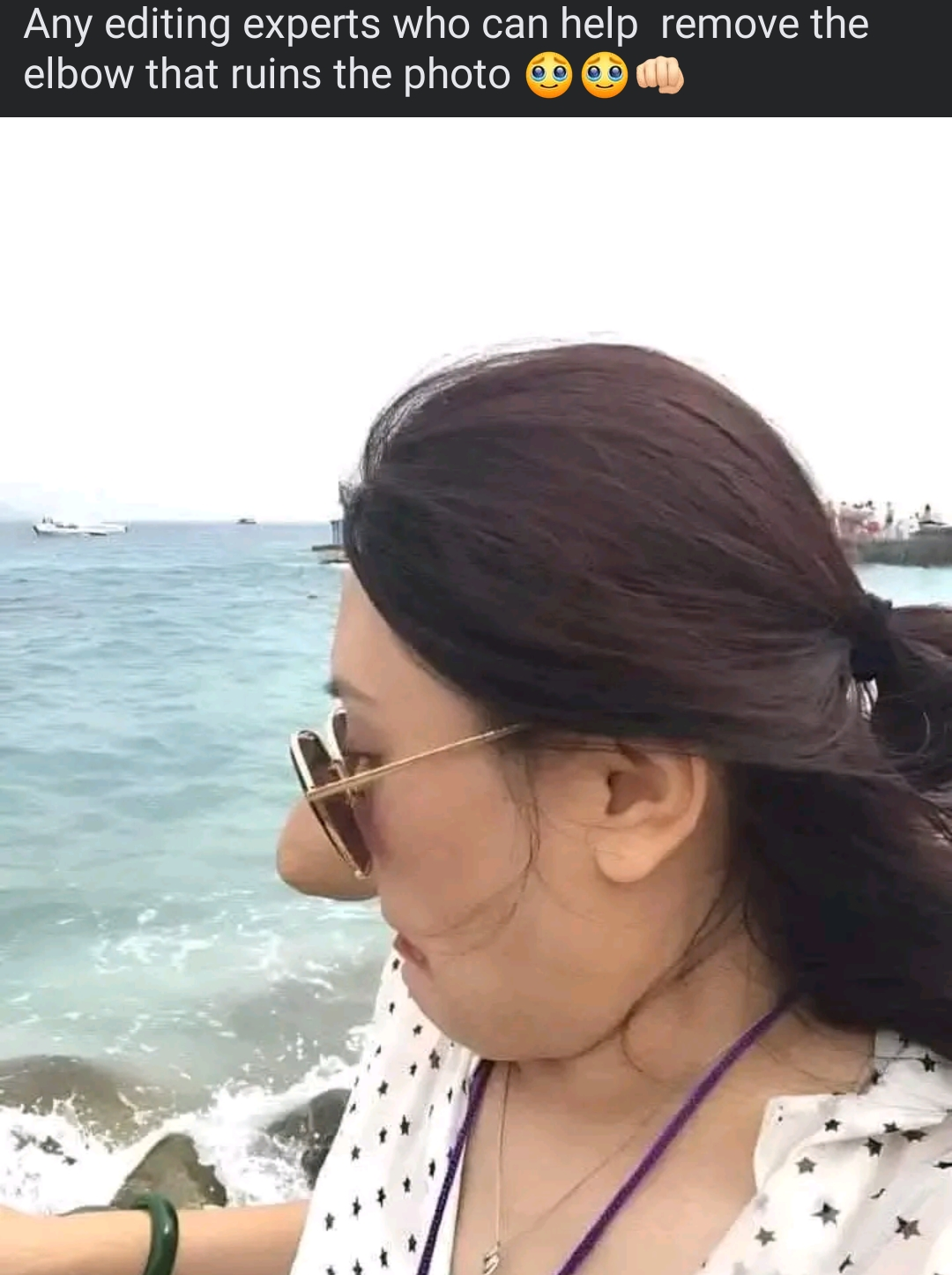 A woman by the sea wearing sunglasses looks at the water. Another person’s elbow, partially visible, appears in the corner, slightly obstructing the scene.
