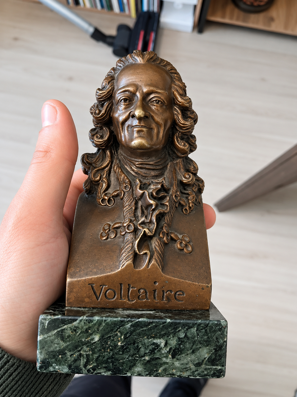 A hand holds a bronze bust of Voltaire with his name engraved on a green marble base. Wooden floor and books are visible in the background.