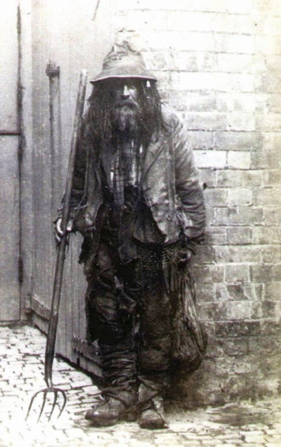 A bearded person in worn, ragged clothes and hat stands on a cobblestone street, holding a pitchfork, with a brick wall and wooden door behind them.