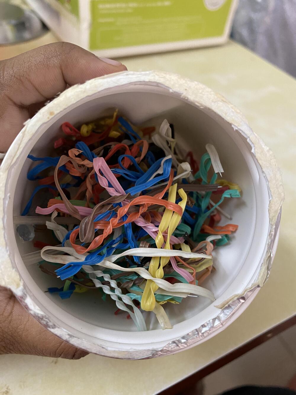 A hand holds a white container filled with assorted colorful rubber bands, some twisted together. The background shows a beige surface and a green and white box.