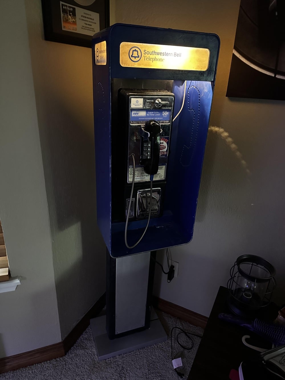 A Southwestern Bell payphone booth stands indoors, illuminated by its sign. The payphone has a corded handset and coin slot. Nearby are a table with items and a window.