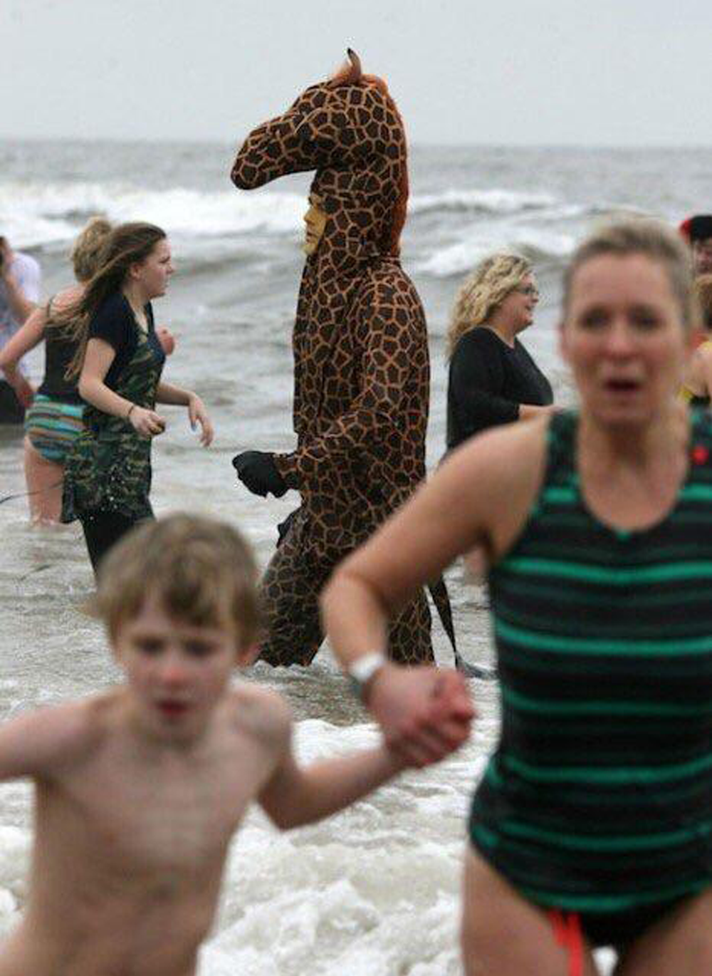 A person in a giraffe costume stands in ocean water among people in swimsuits. Some are running or walking in the waves, and the sky is overcast.