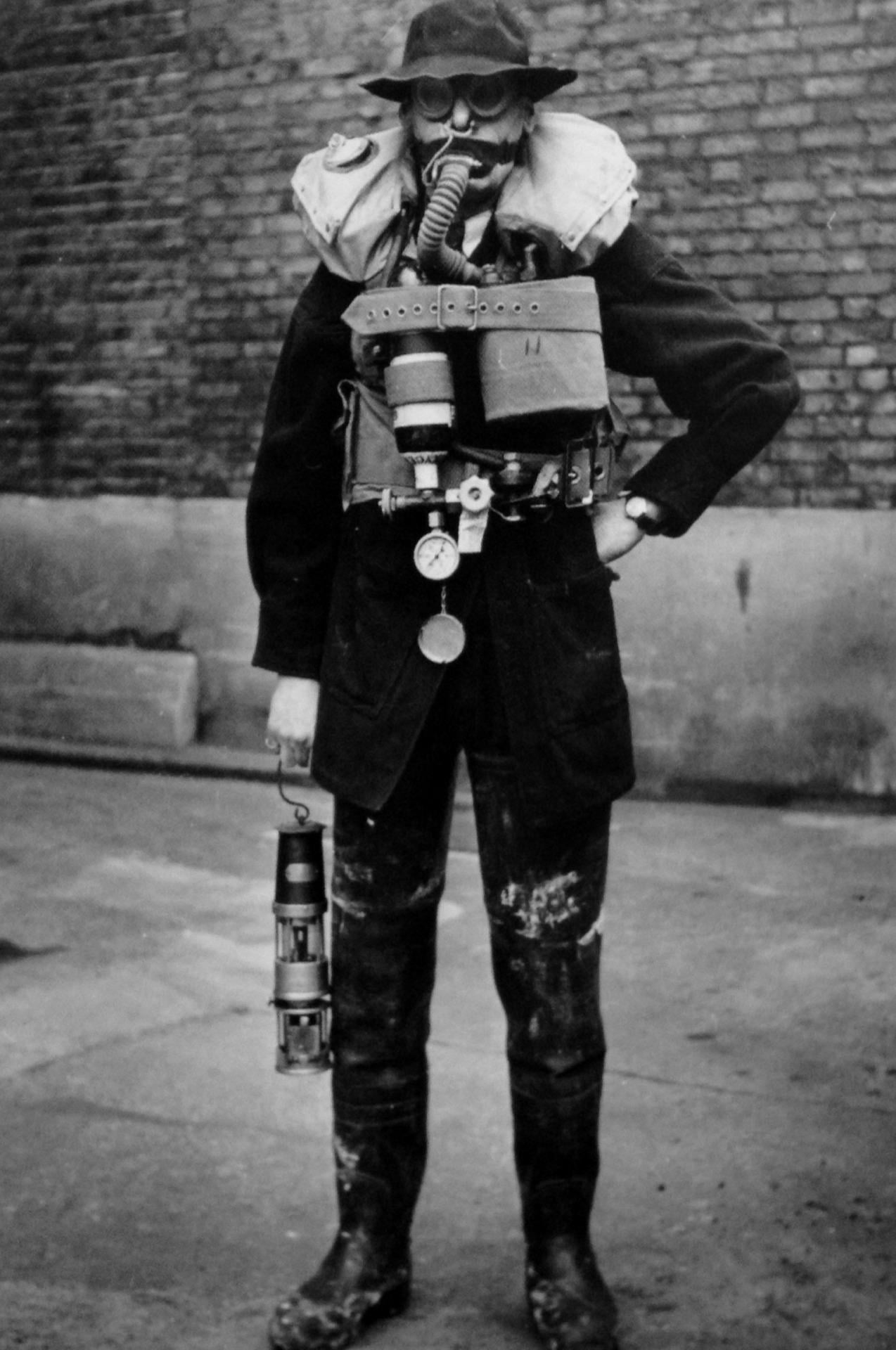 A person wearing protective gear, including a gas mask, heavy boots, and equipment, stands on a street next to a brick wall, holding a lantern in one hand.