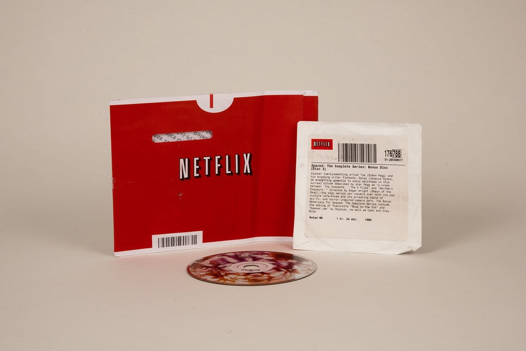 A red Netflix DVD sleeve, a white rental envelope with a barcode and text, and a DVD disc are displayed on a plain background.