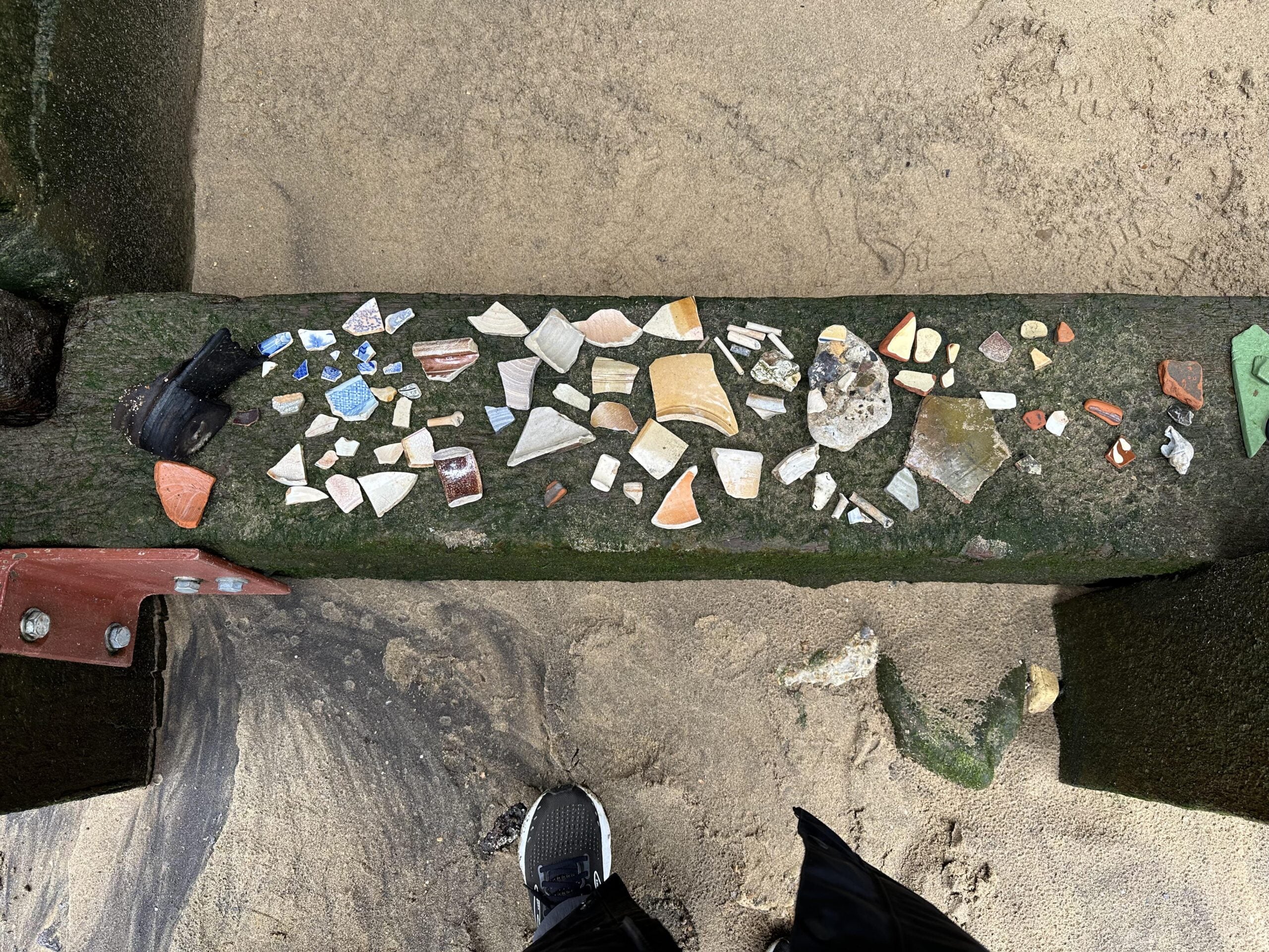 Colorful shards of pottery and glass arranged on a mossy wooden beam at the beach, with sand below and part of a shoe visible at the bottom.