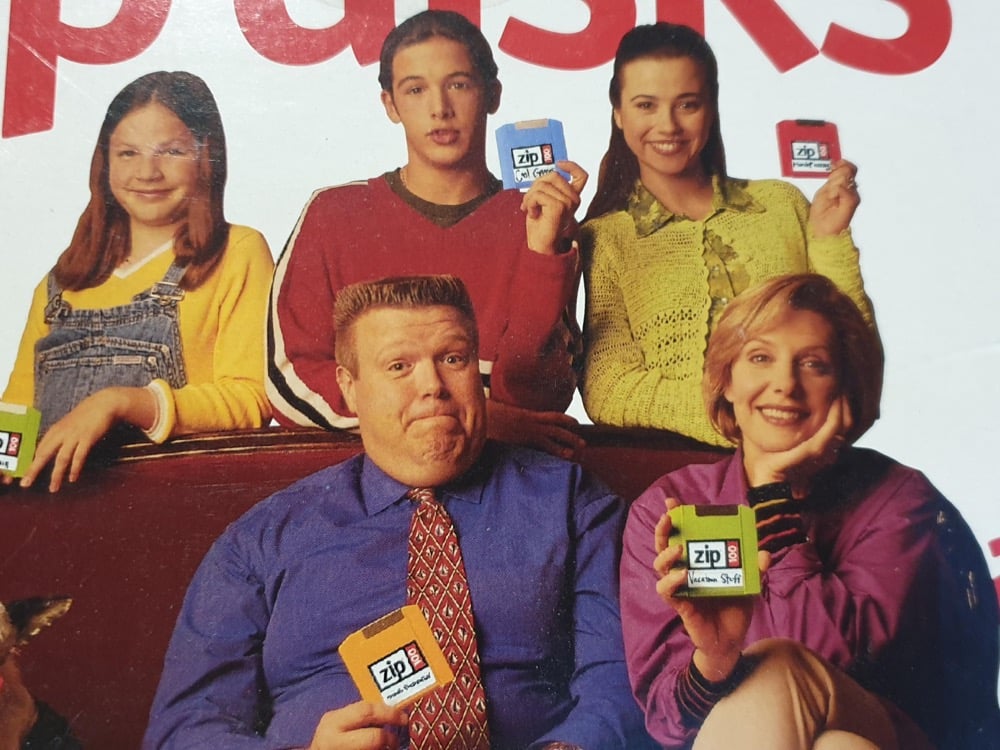A group of five people poses, smiling and holding colorful Zip disks. Three are seated in front; two stand behind. All wear casual 1990s-style clothes, and the background is plain white with red partial text visible at the top.