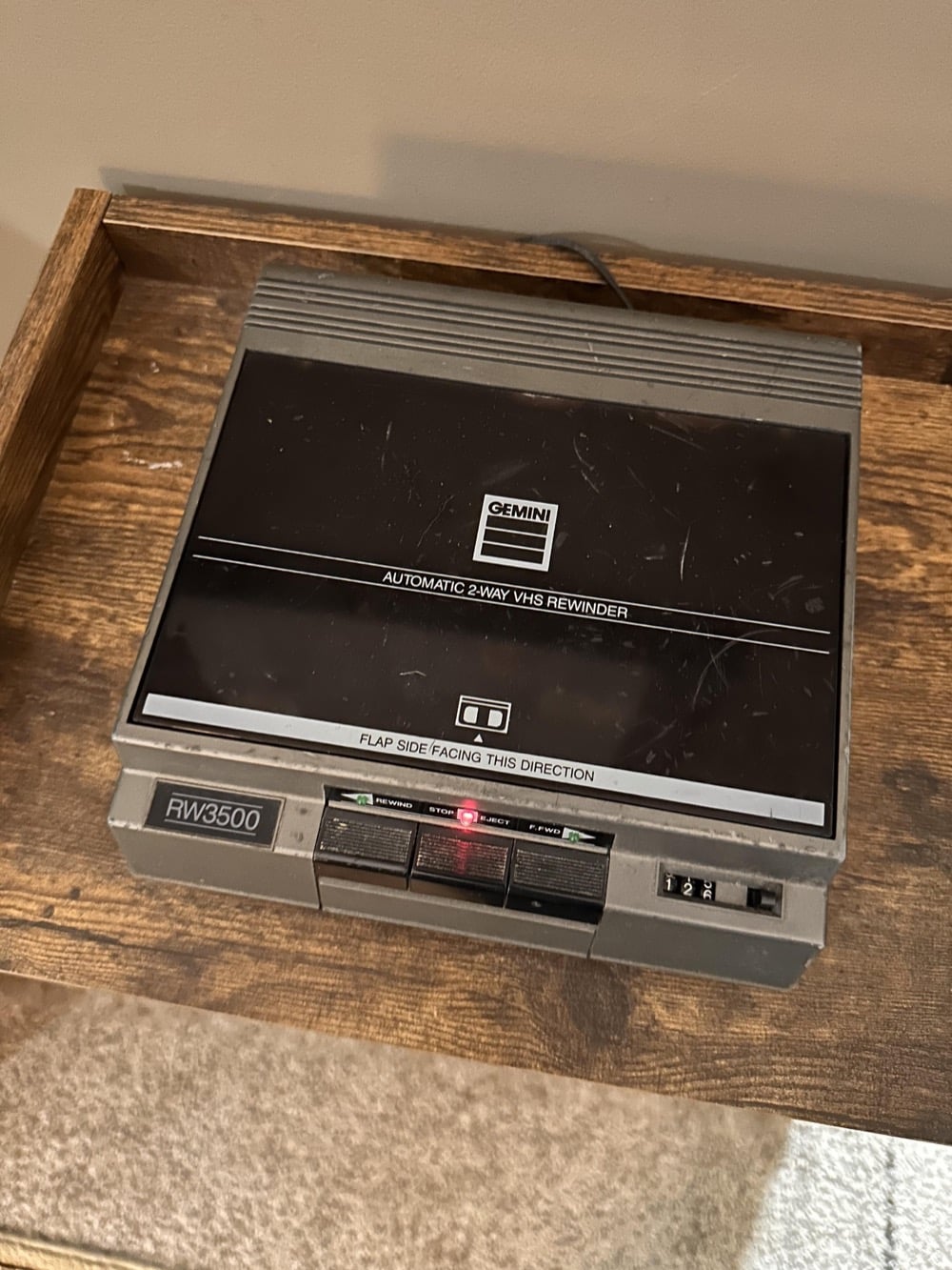 A Gemini RW3500 automatic 2-way VHS rewinder sits on a wooden shelf, plugged in, with a red indicator light illuminated on the front panel.