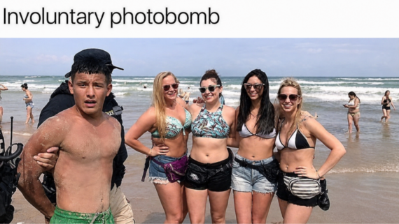 A group of four smiling women in swimsuits pose on a beach while a surprised young man in swim trunks unintentionally appears in front of them. Other beachgoers are visible in the background.