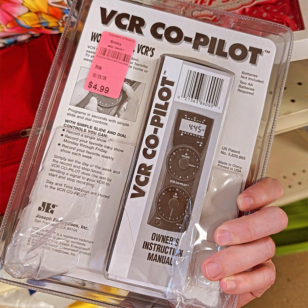 Person holding a packaged VCR CO-PILOT remote, with owner’s manual visible inside. A pink price sticker shows $4.99. The package includes product information and instructions.
