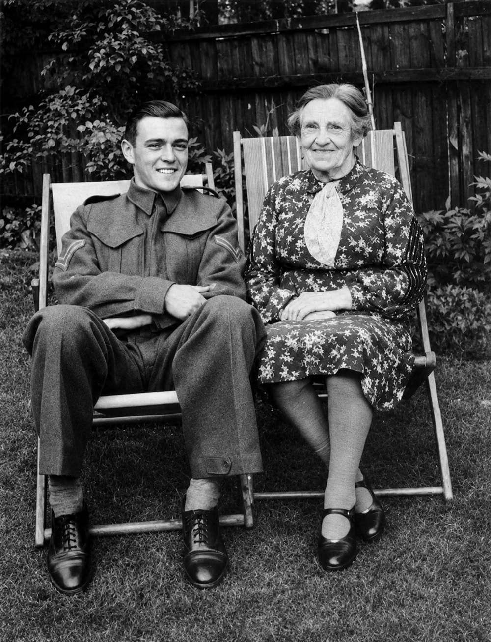 A young man in a military uniform and an elderly woman in a patterned dress sit on lawn chairs in a garden, smiling at the camera.