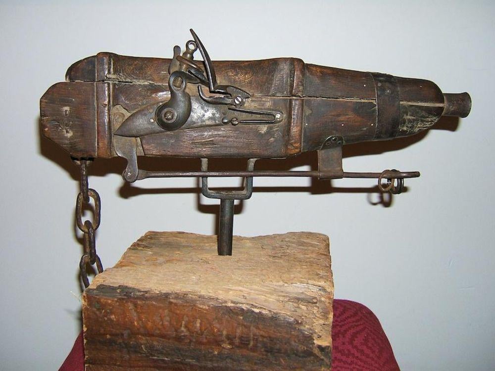 An old, wooden, and metal hand cannon with a flintlock mechanism is mounted on a wooden base, with a chain attached to its side.