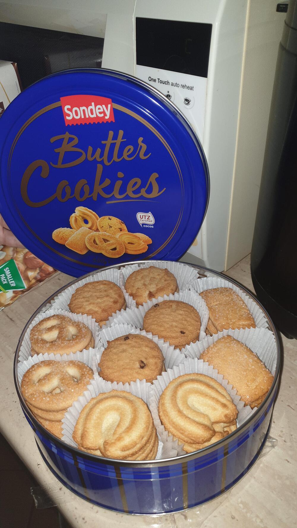 Open blue tin labeled “Sondey Butter Cookies” on a counter, showing assorted butter cookies inside white paper cups. A microwave and other kitchen items are in the background.