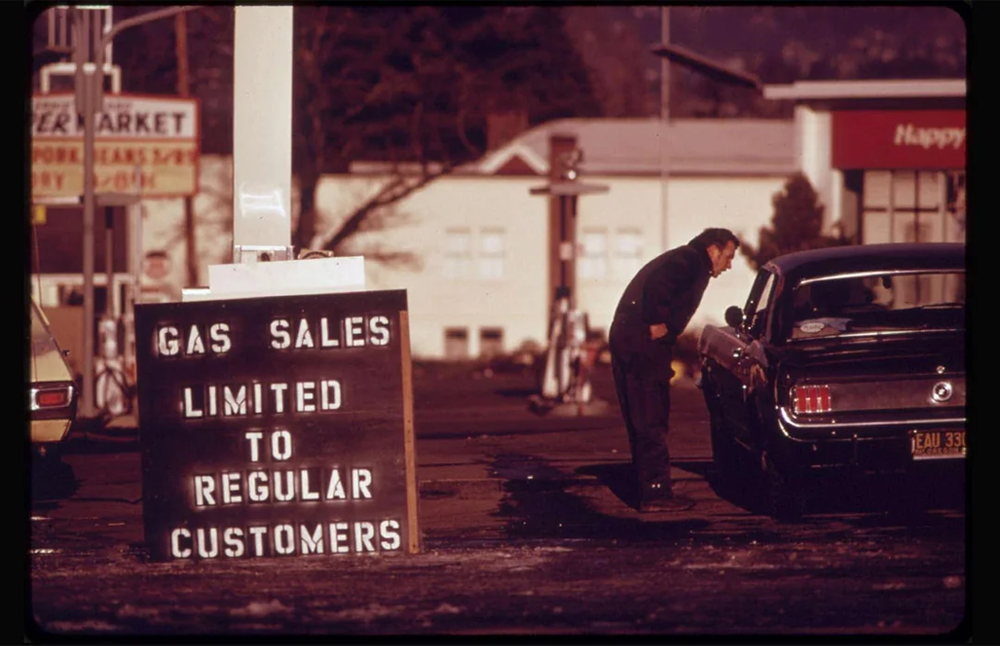 A man leans into a black car at a gas station. A large sign reads, “Gas sales limited to regular customers.” Other cars and buildings are visible in the background.