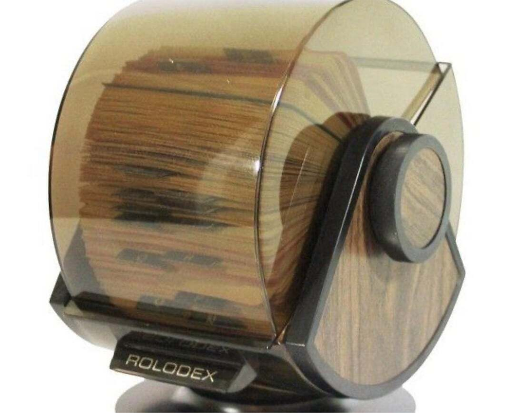 A brown and black Rolodex card file organizer holds many index cards, covered by a tinted plastic lid, on a desk.