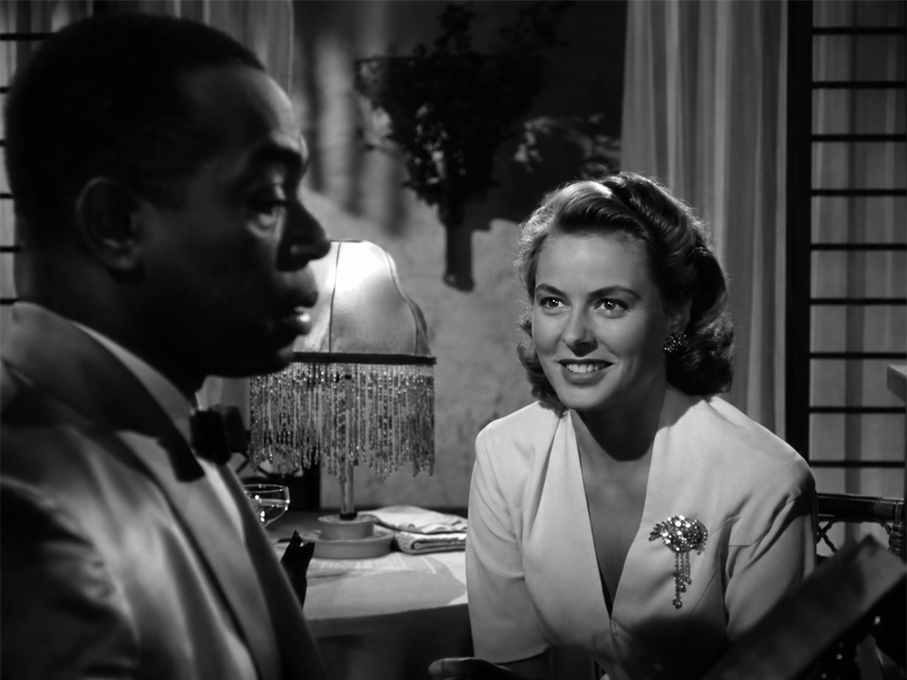 A woman in a white dress with a brooch smiles at a man in a suit. They are seated at a table with a fringed lamp in a softly lit room. The scene is in black and white.