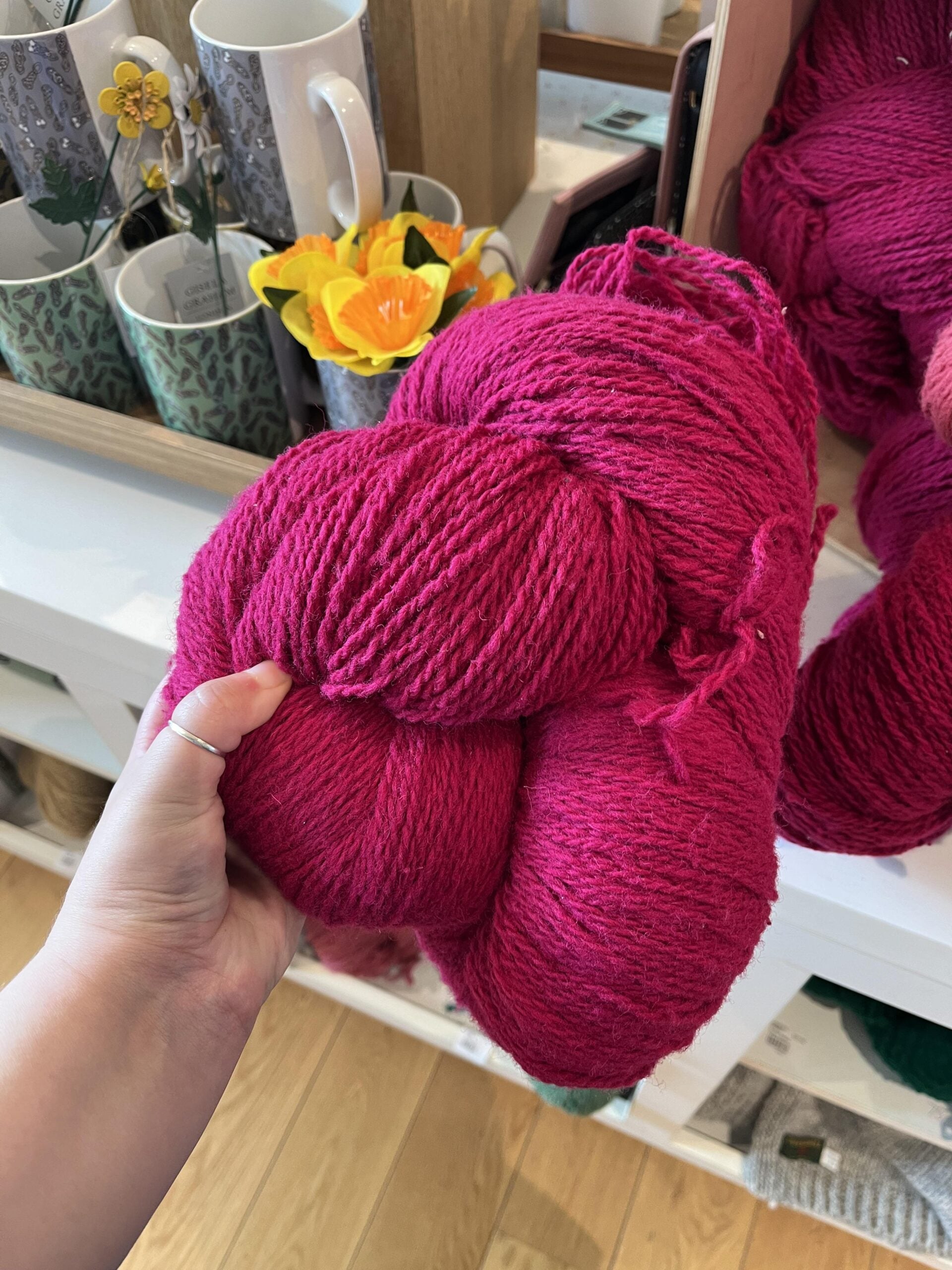 A hand holds a large skein of bright pink yarn in front of a shelf with mugs, a mirror, and a small yellow flower arrangement.