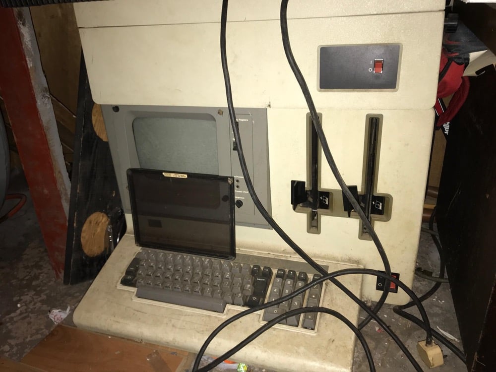 An old, beige vintage computer with a built-in monitor, keyboard, floppy disk drives, and tangled black cables in front on a dusty floor.