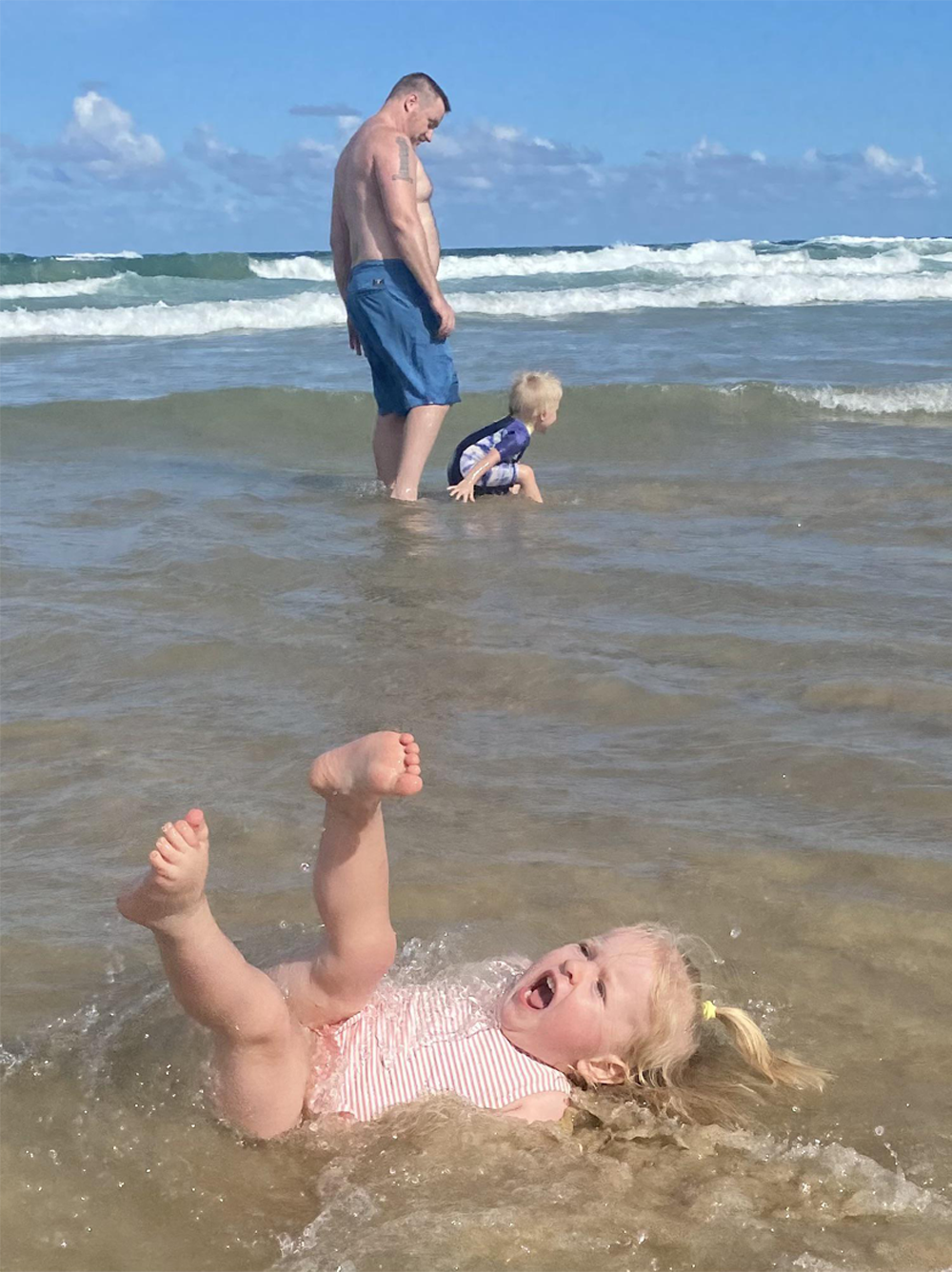 A toddler in a striped swimsuit falls back laughing in shallow ocean water, while a man and another child stand farther away in the waves under a blue sky.