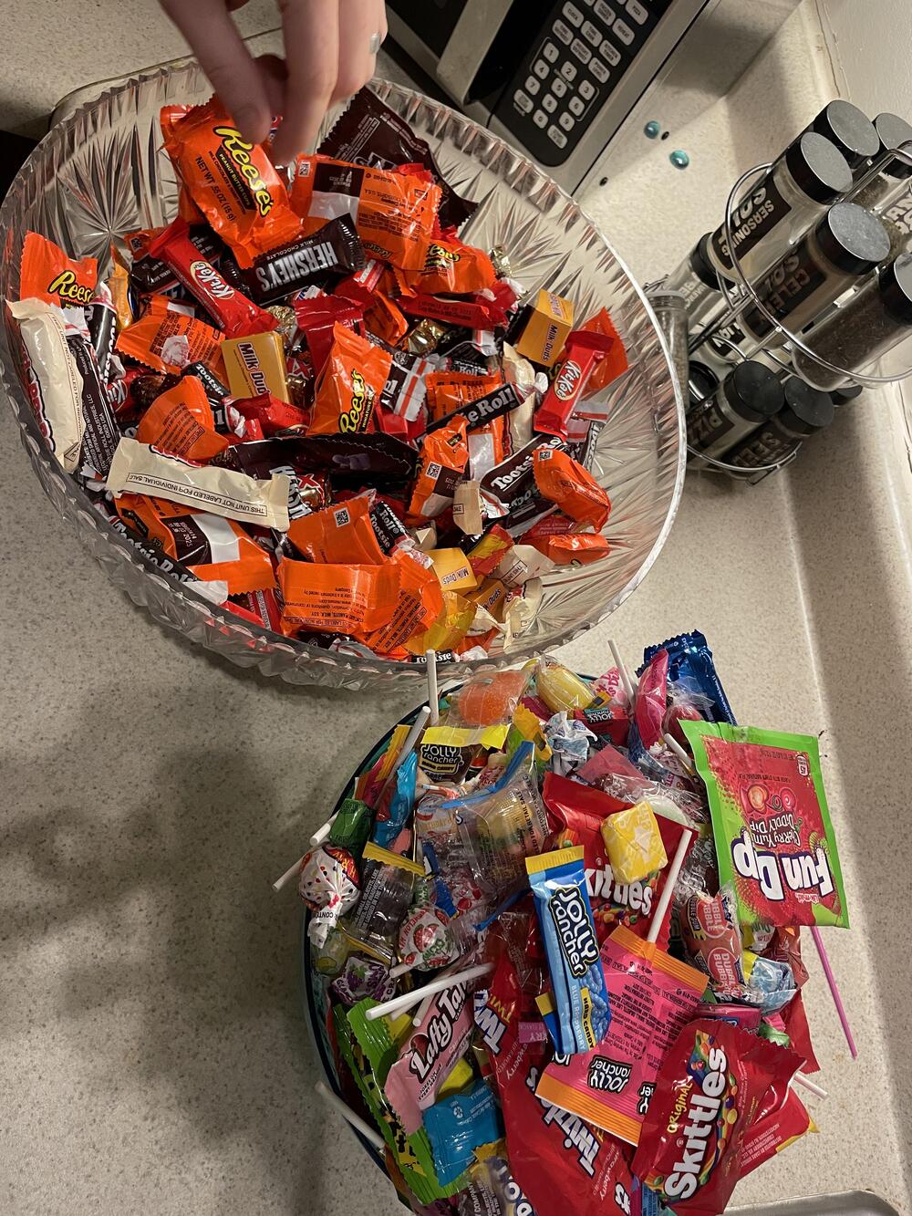 Two bowls of assorted Halloween candy sit on a kitchen counter, with a hand reaching into the top bowl. Candies include Reese’s, Kit Kat, Skittles, Dum Dums, Fun Dip, and more.
