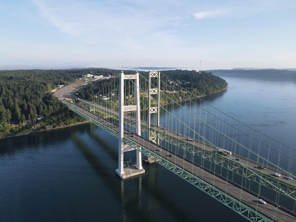 Aerial view of a large suspension bridge crossing over a wide body of water, with vehicles on the bridge and a forested shoreline in the background.