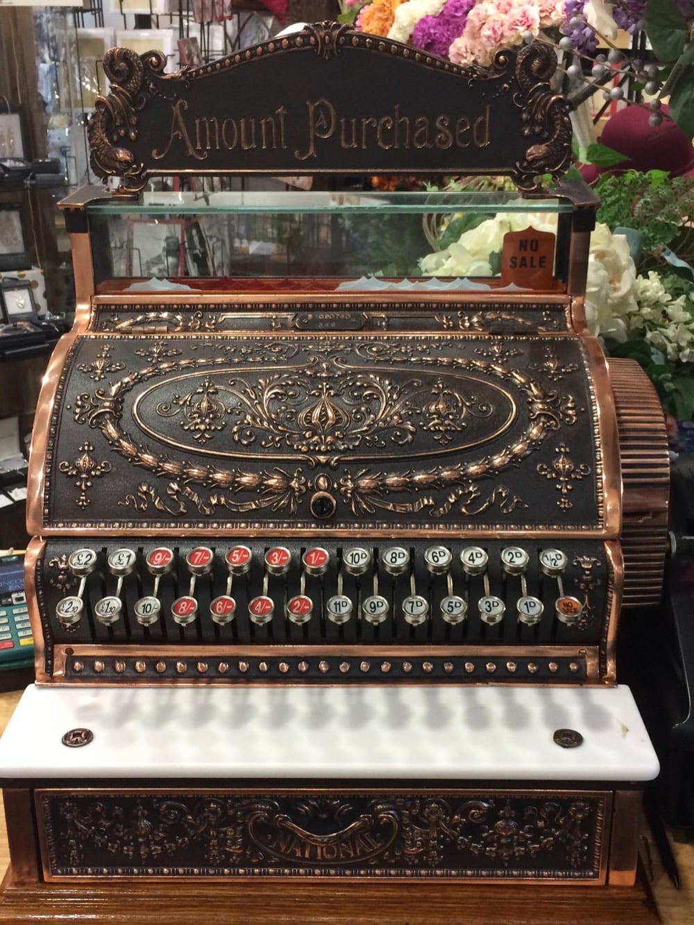 Ornate vintage cash register with decorative floral patterns, labeled “Amount Purchased” on top. Rows of numbered keys and a white base are visible, surrounded by flowers and shop items in the background.
