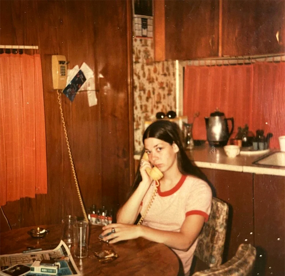 A woman with long dark hair sits at a kitchen table, holding a yellow corded phone to her ear and looking at the camera. The room has wood panel walls, orange blinds, and 1970s-era decor.