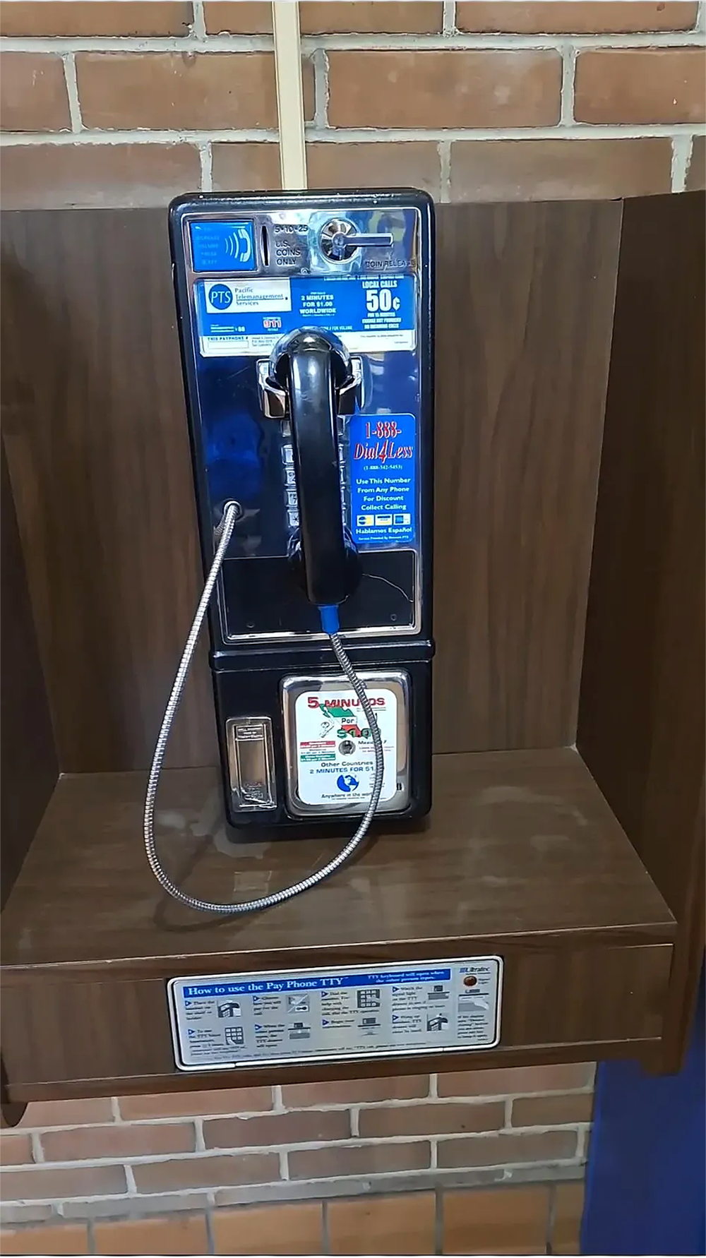 A black, wall-mounted payphone with a corded handset and coin slot, set against a brown wooden panel and a brick wall background. Various instructions and stickers are visible on the front.