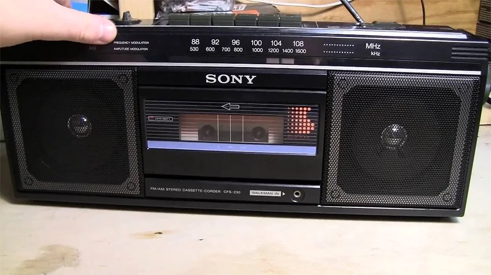 A black Sony boombox with dual speakers, a cassette tape window, and FM/AM radio tuning dial sits on a light wooden surface. A hand is adjusting the controls on the top left corner.