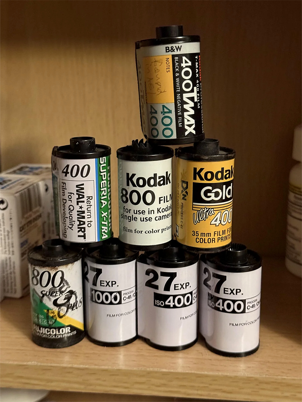 Seven film canisters, including Kodak Gold, Kodak 800, Superia X-tra 400, and others, are stacked on a wooden shelf, with some labeled as 27 exposures and ISO 400. Other shelf items are partially visible in the background.