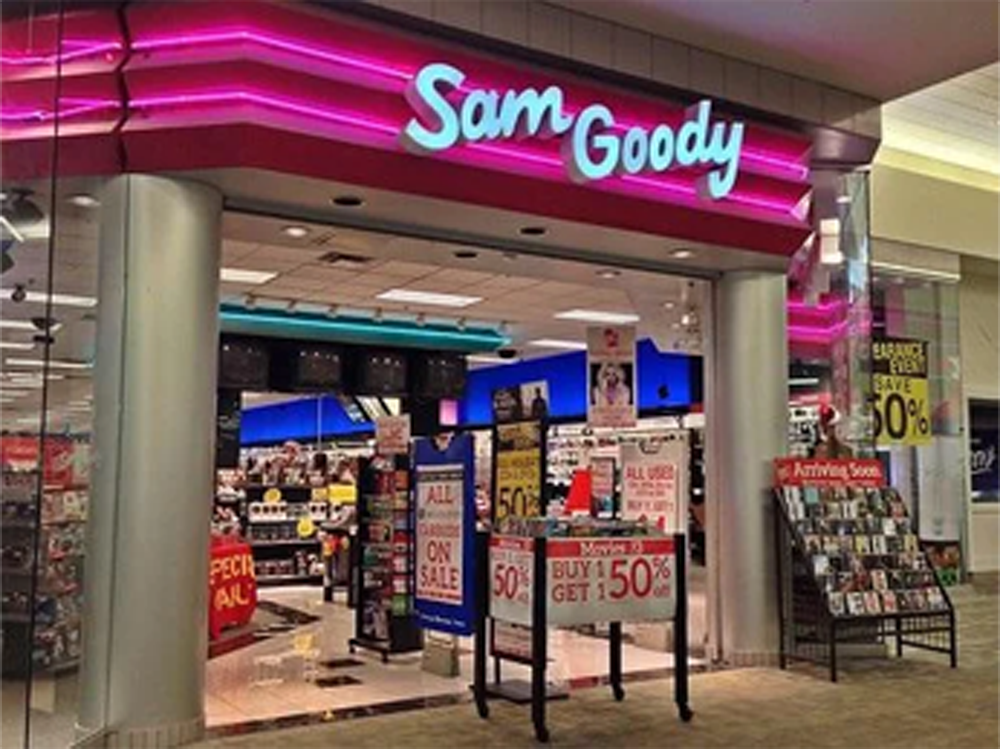 Sam Goody store entrance with bright pink neon sign, sale posters in windows, music and movies displayed inside, and “Buy 1 Get 1 50% Off” and “Special All On Sale” signs visible.