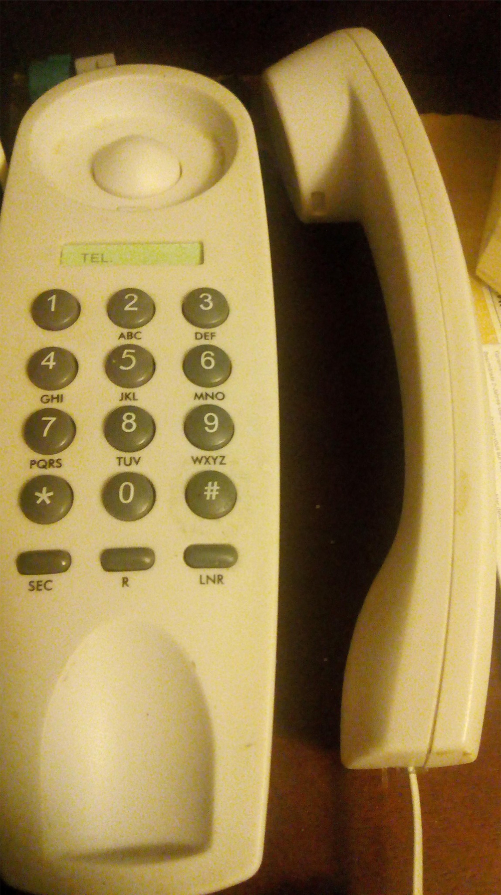 A white landline telephone with the handset off the receiver rests on a dark surface. The phone has a numeric keypad and a small display above the buttons.