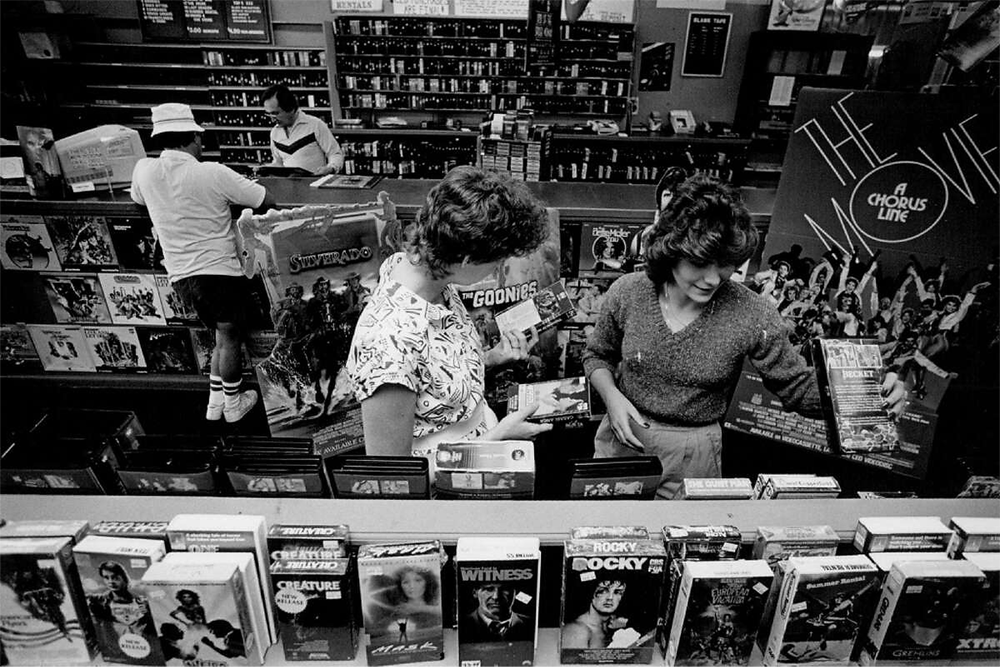 Black-and-white photo of people browsing in a video and music store. Posters, vinyl records, and VHS tapes like “Goonies” and “Rocky” are visible. Two women look at records while another person checks shelves.