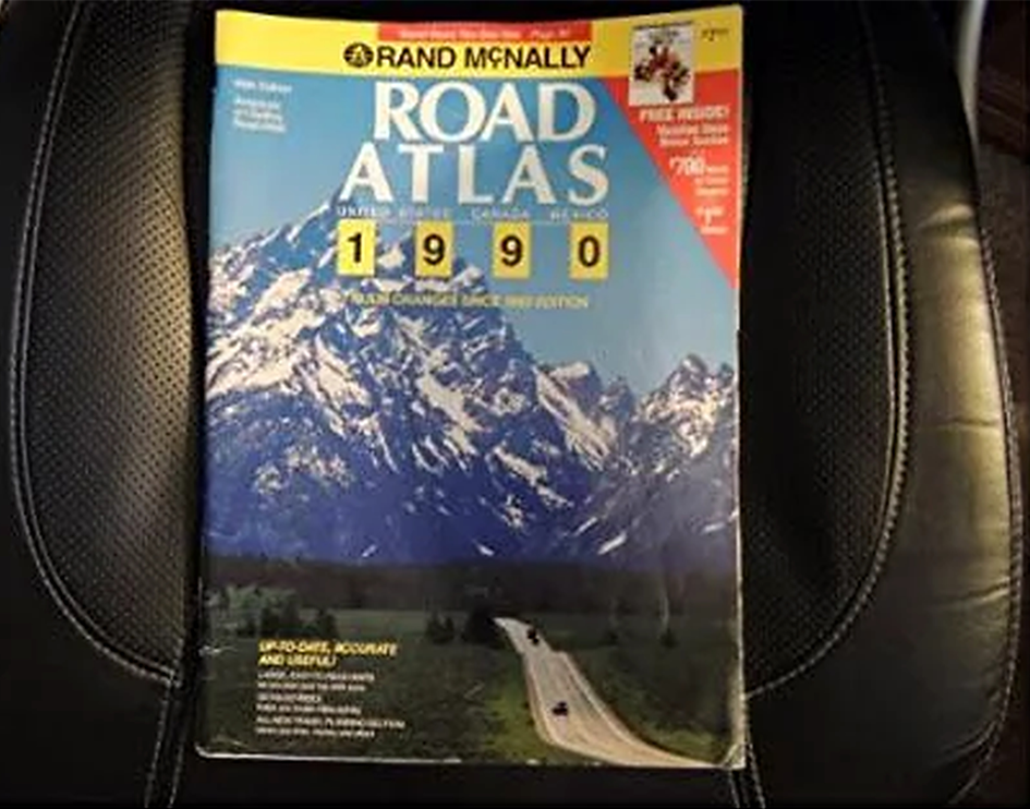 A 1990 Rand McNally Road Atlas rests on a black leather car seat. The cover shows a scenic mountain landscape above a winding highway with cars.
