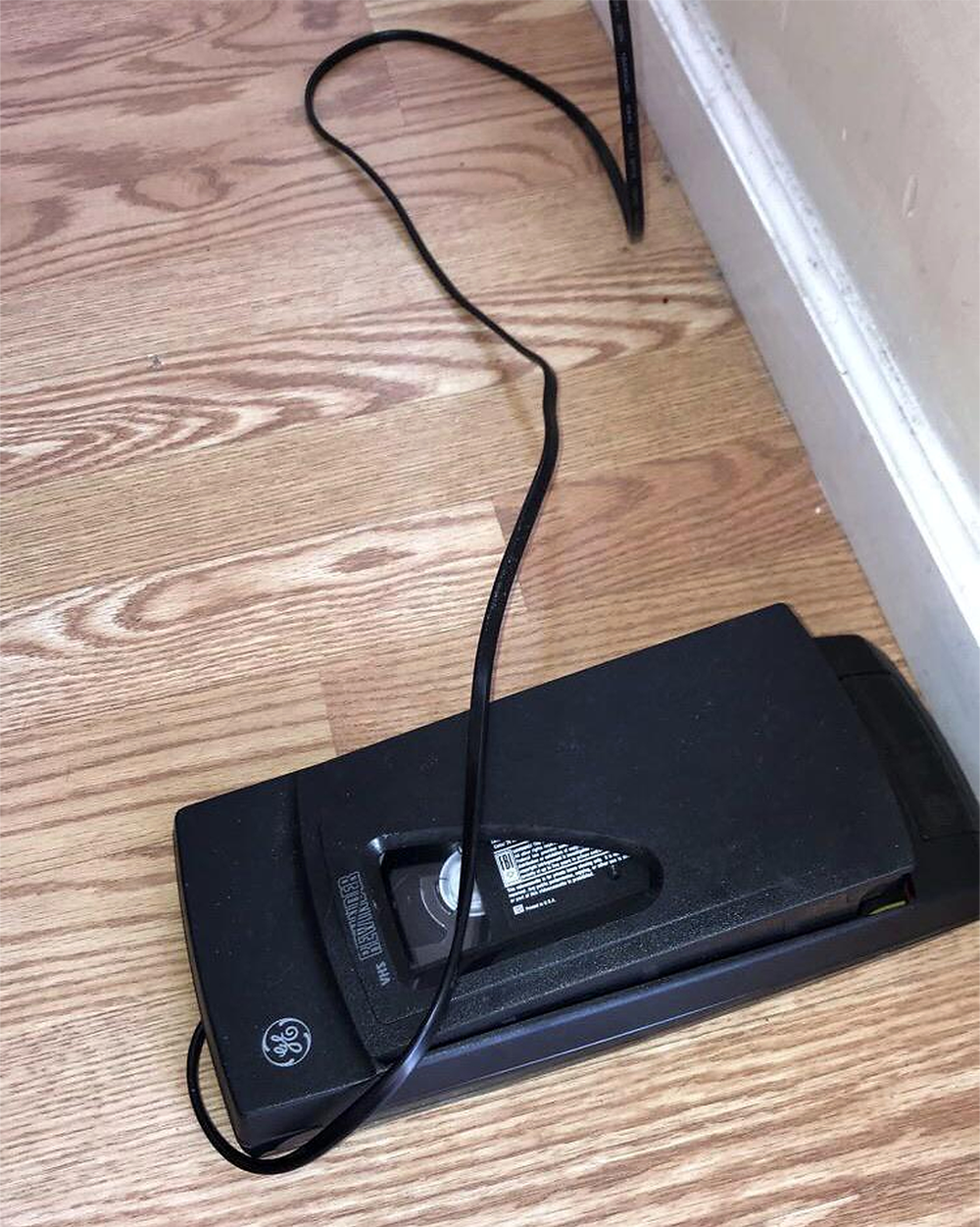 A black GE iron lies face down on a wooden floor near a white baseboard. Its electric cord is loosely coiled on the floor.