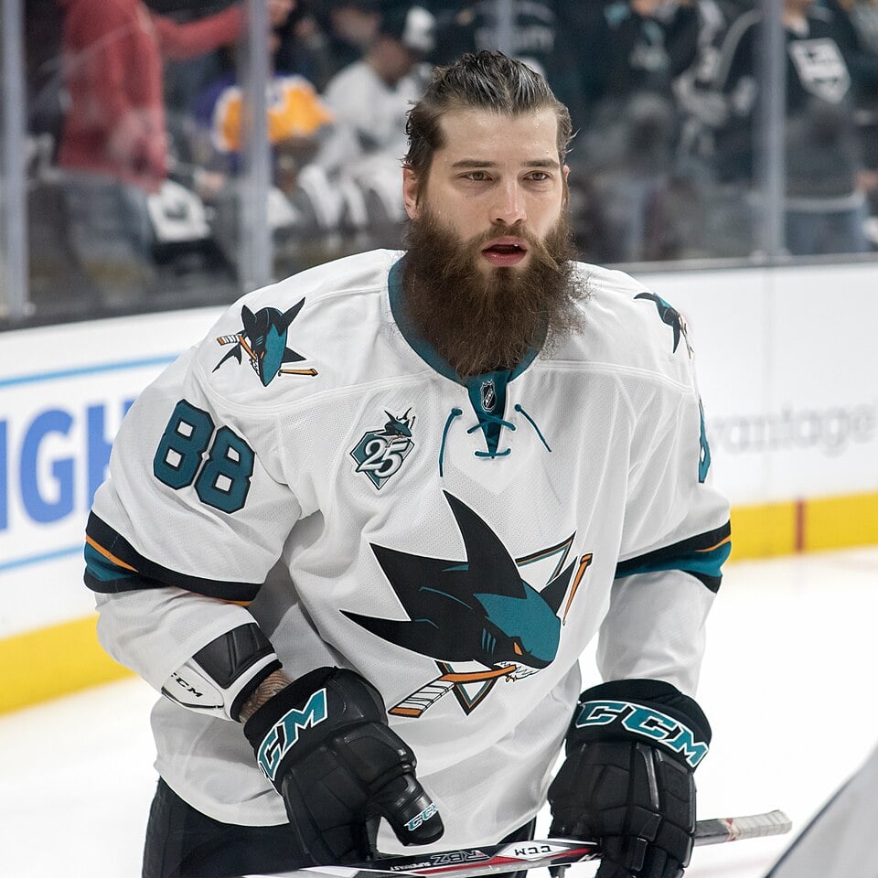 A hockey player in a white San Jose Sharks jersey with the number 88 and a large beard stands on the ice, holding a hockey stick. The background shows blurred spectators and other players.