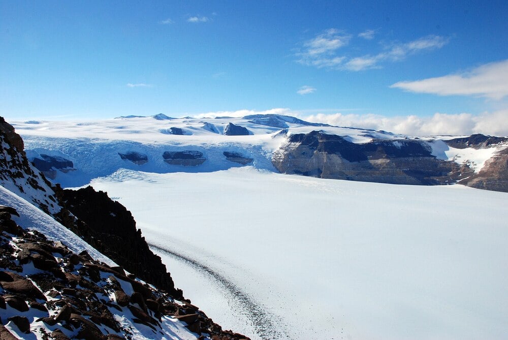 Snow-covered mountains and a vast glacier under a clear blue sky, with rocky terrain in the foreground and patches of ice and snow visible across the landscape.