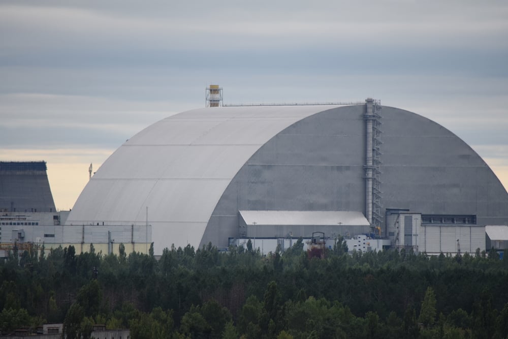 A large, arched metal structure stands behind trees under a cloudy sky; this is the New Safe Confinement at the Chernobyl Nuclear Power Plant in Ukraine.