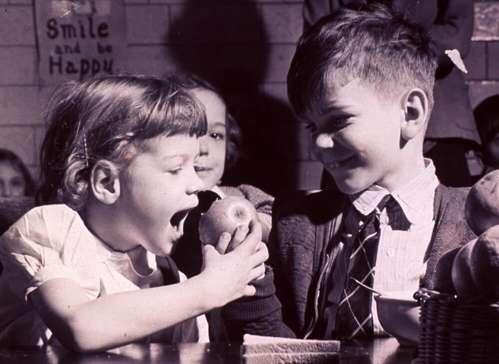 A young girl excitedly opens her mouth to eat an apple while a smiling boy holds it out to her. Other children watch in the background. A sign on the wall reads, “Smile and be Happy.”