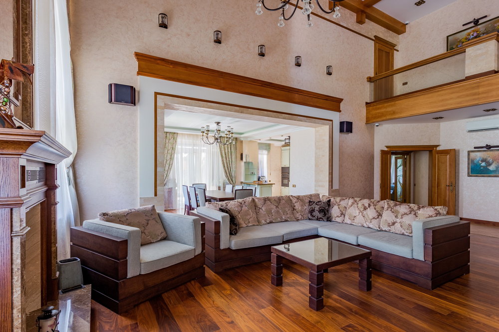 Modern living room with a large L-shaped sofa, glass coffee table, wooden floors, and high ceilings. Open view to a bright kitchen and dining area with large windows and light curtains.