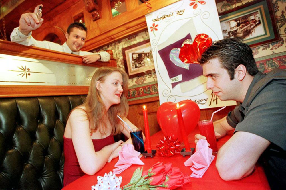 A couple sits at a decorated restaurant table, holding hands with drinks and a candle between them. A waiter stands behind, smiling and holding a figurine. A Valentine’s Day card and decorations are on the table.