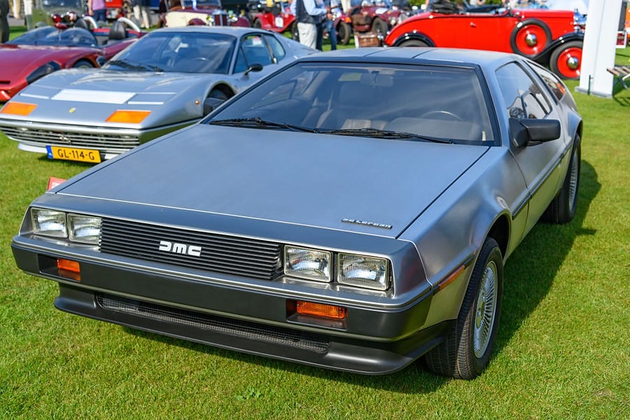 A silver DeLorean DMC-12 sports car is parked on grass at a car show, surrounded by other vintage vehicles in the background.