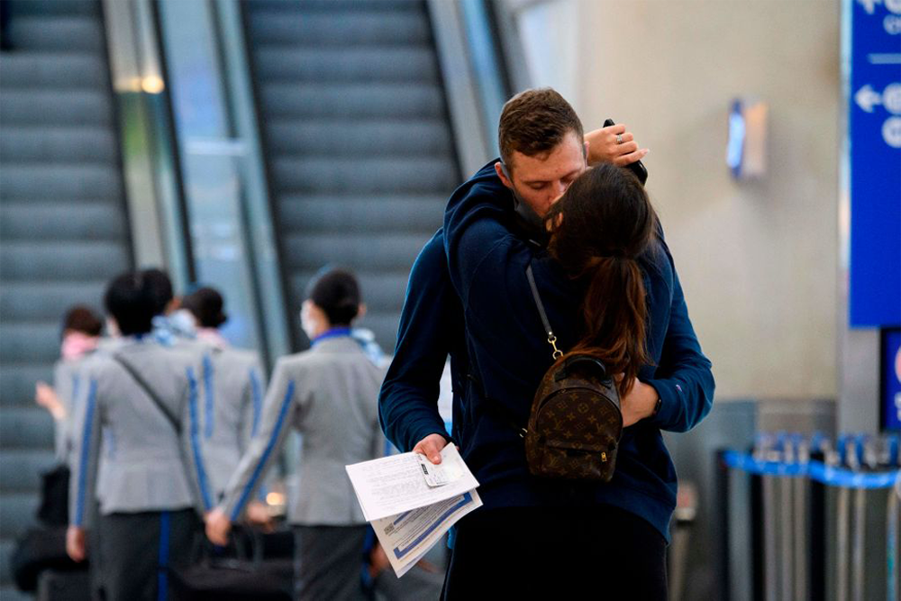 A couple embraces and kisses in an airport terminal, while holding travel documents. Escalators and airline staff in uniforms are visible in the background.