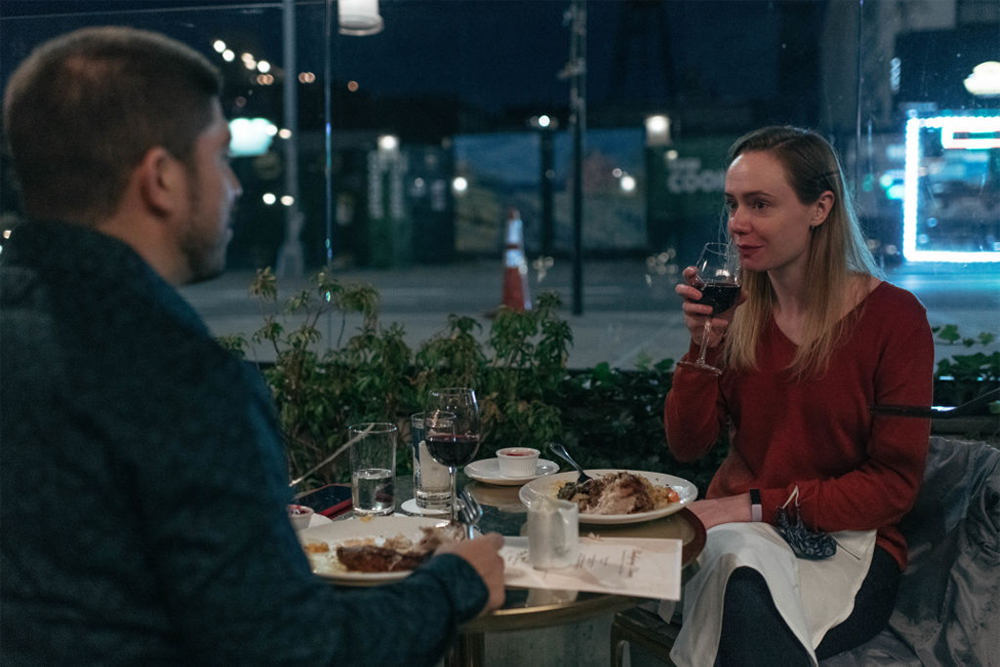 Two people sit at an outdoor restaurant table at night, eating dinner and drinking wine. The woman is sipping from her glass while the man, seen from behind, faces her. Urban lights and street scene are visible in the background.