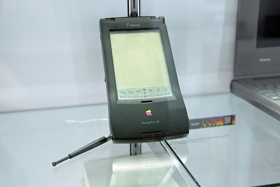 A vintage Apple Newton MessagePad 120 PDA with a stylus resting against it, displayed in a glass case. The device features a small black-and-white screen and an old multicolor Apple logo.