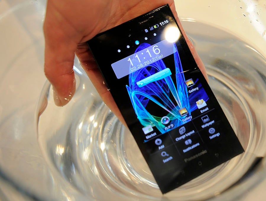 A hand submerges a smartphone in a clear bowl of water, with the phone screen lit and showing app icons and the time 11:16.