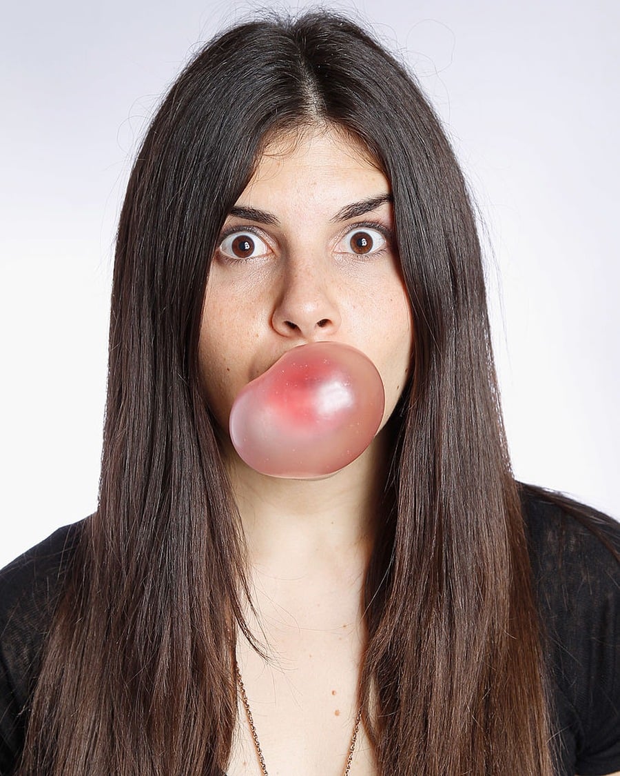 A woman with long brown hair and wide eyes blows a pink bubble with chewing gum, looking surprised against a plain white background.